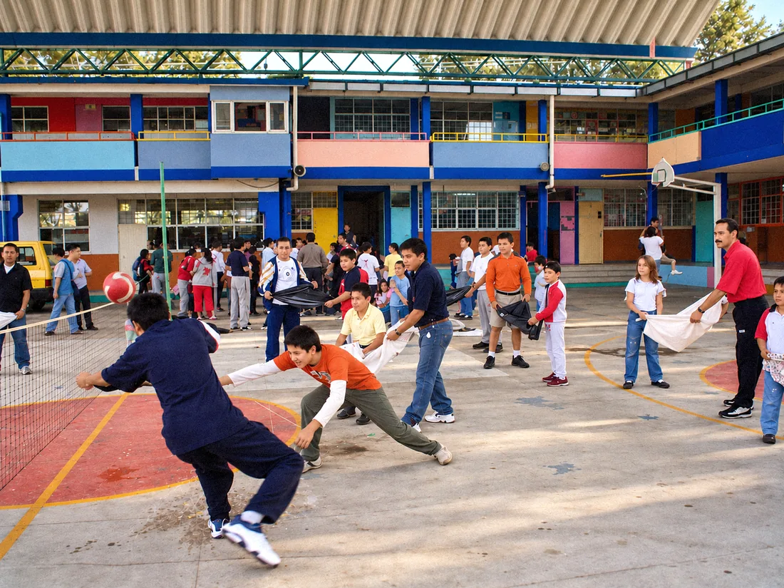 Estudiantes jugando en la cancha frente al edificio de dos plantas, con fachada azul, rosa y naranja y tablero de basquetbol